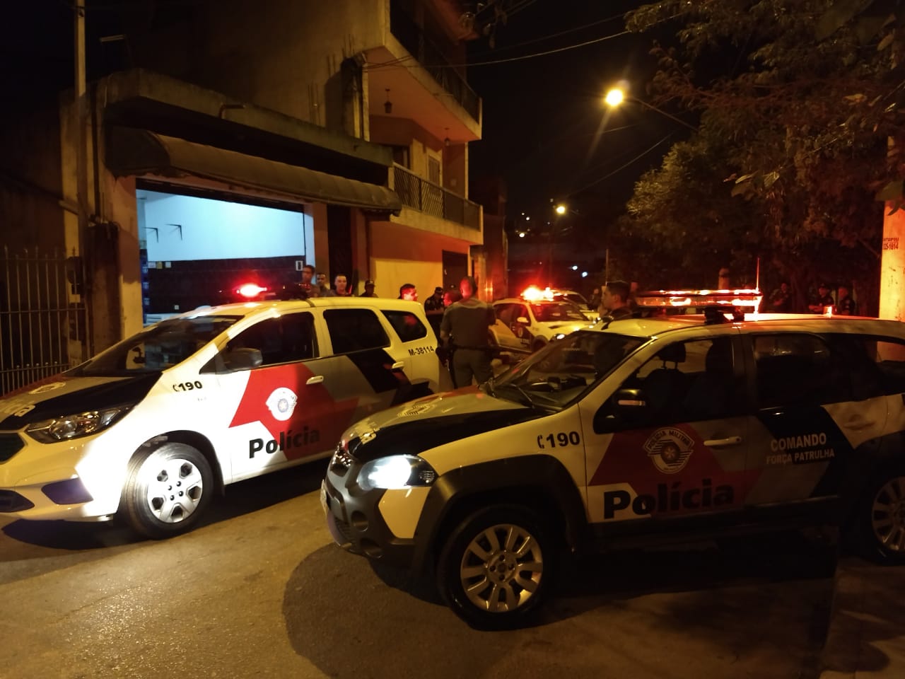 viaturas da Polícia Militar, com luzes de alerta acesas bloqueiam rua, em frente ao que deve ser um bar, que está com a porta de ferro aberta e iluminado. Um policial está indo em direção à porta, de costas, entre os dois carros policiais. Ao fundo, alguns homens observam. Só está visível a cabeçadeles, acima do teto da viatura. Um guarda civil metropolitano está em pé, também ao fundo, ao lado de um carro da GCM. No outro lado da rua, imagem desfocada de três policiais.